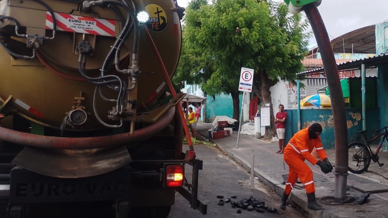 a imagem mostra a traseira de um caminhão limpa fossa e um operário colocando uma mangueira em um bueiro